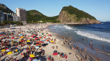 Uma animada cena de praia mostra guarda-sóis coloridos, banhistas e pessoas brincando nas ondas. Montanhas verdes e um céu azul criam uma atmosfera de verão vibrante e relaxante.
