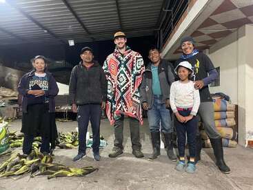 Six people and one child stand together, smiling under a metal roof. Corn is scattered on the ground. Everyone appears happy and relaxed, likely in a rural setting.
