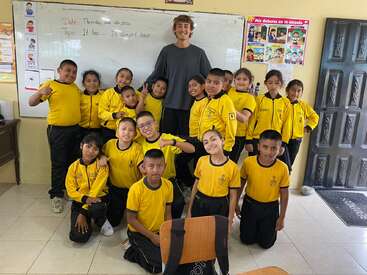 A smiling teacher stands with a group of cheerful students in yellow uniforms inside a classroom. They pose together in front of a whiteboard and posters.