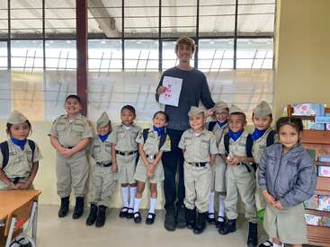 A group of young children in uniform stand with an adult holding a drawing. They are smiling inside a classroom with bright windows in the background.