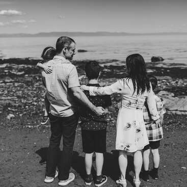 Une famille de quatre personnes se tient sur une plage rocheuse, face à l'océan. Les parents embrassent doucement leurs enfants. Ce moment est capturé en noir et blanc.