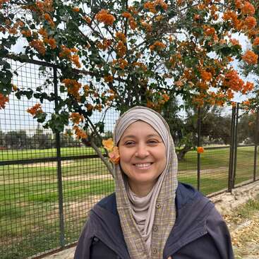 Eine lächelnde Frau mit Kopftuch steht vor einem Zaun, hinter ihr hängen orangefarbene Blumen an einem Baum und sie hat sich eine ans Ohr gesteckt.