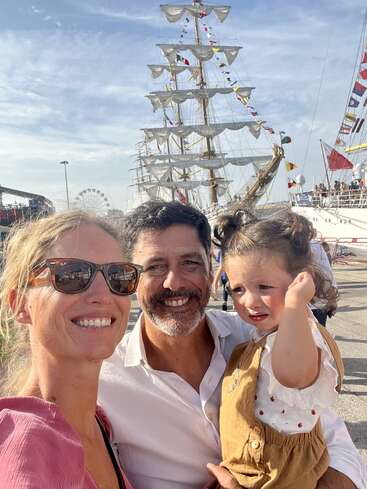 A imagem mostra uma família de três pessoas posando em frente a um grande veleiro, com a mulher à esquerda, o homem no meio e a criança à direita.