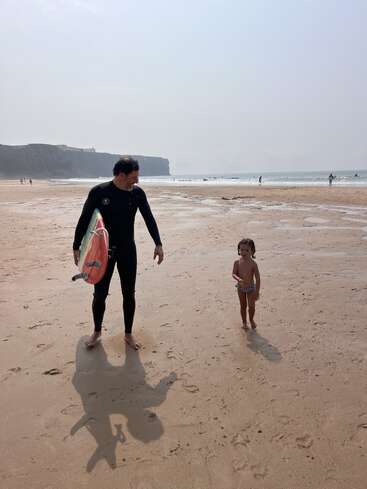 A imagem mostra um homem e uma criança em uma praia, com o homem segurando uma prancha de surfe e usando uma roupa de mergulho, enquanto a criança está vestida com roupas de banho.