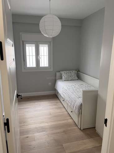 A imagem mostra um quarto pequeno e aconchegante com um sofá-cama, uma janela e uma porta, com um esquema de cores branco e cinza e piso de madeira clara.