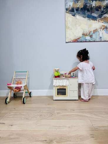 A imagem mostra uma menina brincando com uma cozinha de brinquedo e uma boneca em uma sala com piso de madeira clara e uma parede branca.
