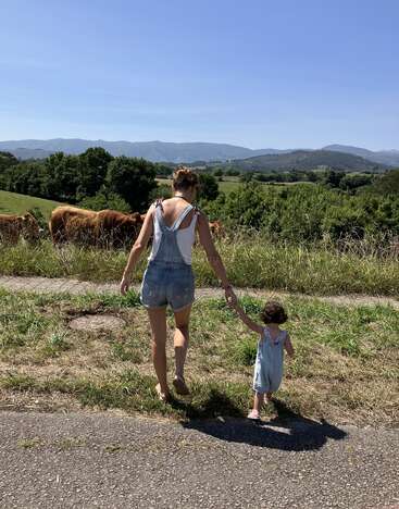 A imagem mostra uma mulher e uma criança caminhando em uma estrada, com um rebanho de vacas ao fundo, em uma paisagem rural pitoresca.