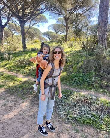 A imagem mostra uma mulher carregando uma criança em uma mochila, andando em um caminho de terra em uma área arborizada, com a criança segurando um bicho de pelúcia.