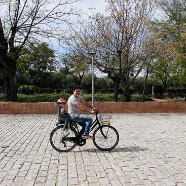 A imagem mostra um homem andando de bicicleta com uma criança em uma cadeirinha, em um cenário de árvores e uma parede de tijolos.