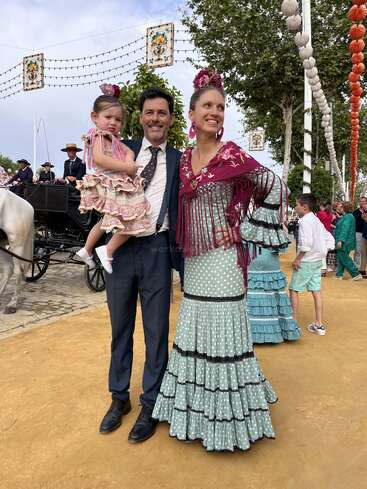 A imagem mostra uma família de três pessoas vestidas com trajes tradicionais espanhóis, posando para uma foto em um evento ao ar livre. A mulher usa um vestido longo com um xale.