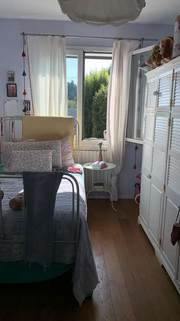 Une chambre confortable avec un lit simple, des oreillers fleuris, des rideaux blancs, un plancher en bois, une bibliothèque, une armoire, des animaux en peluche, une table d'appoint avec des livres et une fenêtre ensoleillée.