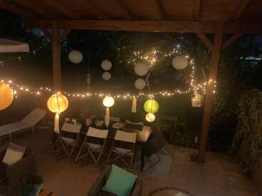 Un patio extérieur confortable est décoré de guirlandes lumineuses, de lanternes colorées et de globes blancs suspendus. Il y a une longue table à manger avec des chaises disposées en dessous.