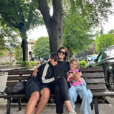 Une femme et deux enfants sont assis sur un banc public et sourient. Ils sont entourés d'arbres verts, de voitures garées et d'un environnement urbain paisible.