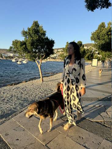 A woman in a patterned outfit walks her dog along a seaside promenade, with trees, boats, and hills in the background, basking in golden sunlight.