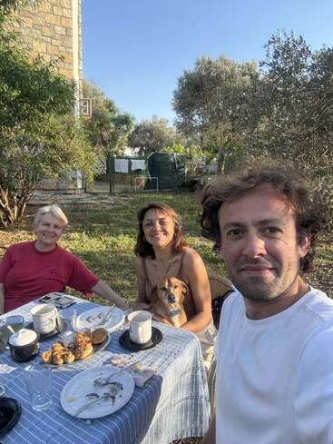 Three people and a dog enjoy an outdoor meal at a table, surrounded by greenery and trees, smiling and relaxed on a sunny, clear day.