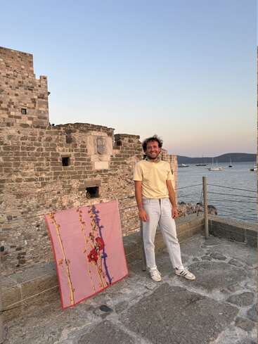 A man in light clothes stands on ancient stone ruins by the sea, smiling. A pink floral cloth is draped nearby. Sailboats float in the background.