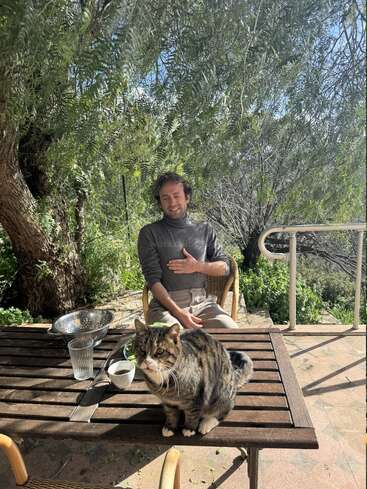 A man sits smiling at an outdoor table under a leafy tree. A tabby cat is sitting on the table. Dappled sunlight completes the peaceful scene.