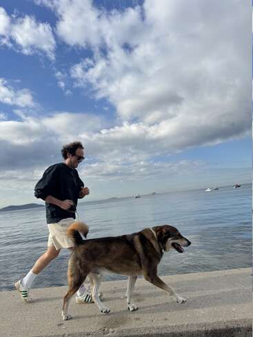 Ein Mann joggt mit seinem großen Hund fröhlich einen Uferweg entlang. Der Himmel ist teilweise bewölkt, und Boote treiben friedlich auf dem ruhigen Meer.