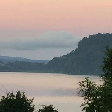 The image depicts a serene lake scene with a mountainous backdrop, surrounded by lush trees and a vibrant sky, evoking a sense of natural beauty and tranquility.