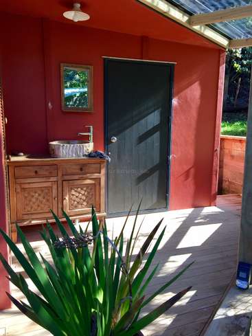 The image depicts the exterior of a small, red building with a wooden door and a sink on a wooden counter, surrounded by a deck and a plant in the foreground.