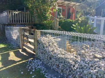 The image depicts a backyard with a wooden deck, a red shed, and a gabion wall made of rocks and wire mesh, surrounded by lush greenery.