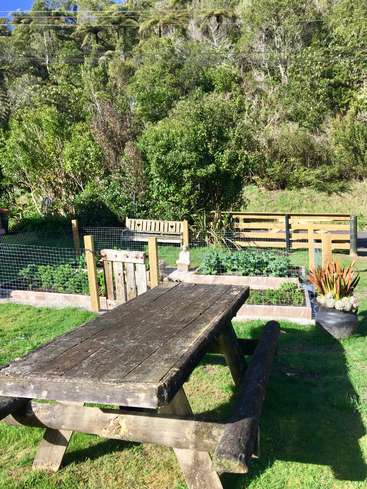 The image depicts a serene outdoor setting, featuring a weathered wooden picnic table, a fenced garden bed, and lush trees, bathed in sunlight.
