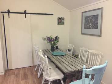 The image depicts a dining room with a rustic wooden table, white chairs, and a sliding barn door, featuring a vase of flowers and a framed picture on the wall.