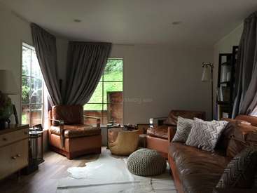 The image depicts a cozy living room with a brown leather couch and chair, a window with gray curtains, and a white rug on a wooden floor.