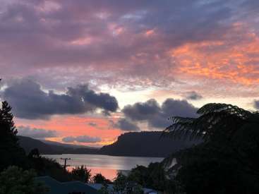 The image depicts a serene sunset over a lake, with a house and trees in the foreground, and a mountainous landscape in the background, under a vibrant pink and purple sky.
