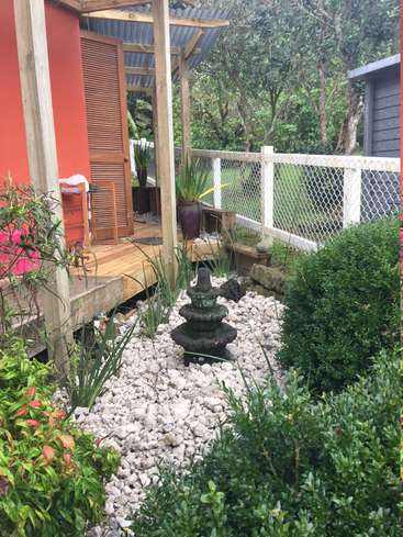 The image depicts a serene outdoor setting, featuring a wooden deck, a small garden with white rocks, and lush greenery, surrounded by a fence and trees.