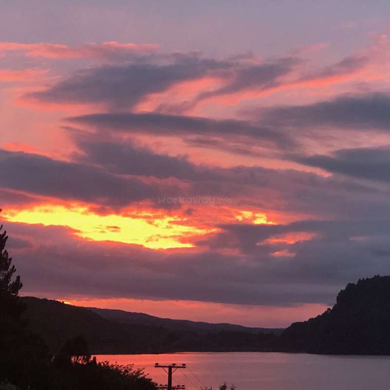 The image depicts a serene sunset over a body of water, with hills and trees in the foreground and a cloudy sky above.