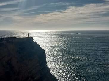 Uma pessoa sozinha está em pé numa falésia, contemplando um oceano vasto e cintilante sob um céu claro com algumas nuvens, transmitindo uma sensação de aventura e tranquilidade.