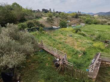 Un paysage champêtre verdoyant et luxuriant, composé d'herbes sauvages, d'arbres, de clôtures rustiques et d'un chemin de terre sinueux, avec au loin des montagnes et des bâtiments agricoles éparpillés sous un ciel nuageux.