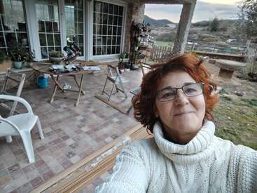 Une femme aux cheveux roux et portant des lunettes sourit pour un selfie sur une terrasse carrelée où se trouvent des tables, des chaises et des plantes, avec en arrière-plan un magnifique paysage rural.