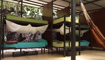 This image shows an open-air dormitory with four bunk beds, mosquito nets, colorful bedding, a hammock, crates for storage, and a lush green background outside.