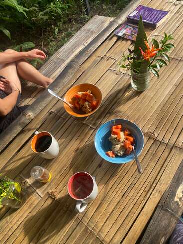A cozy outdoor breakfast scene on a bamboo table, featuring fruit bowls, coffee mugs, honey, fresh flowers, books, and a relaxed person sitting nearby.