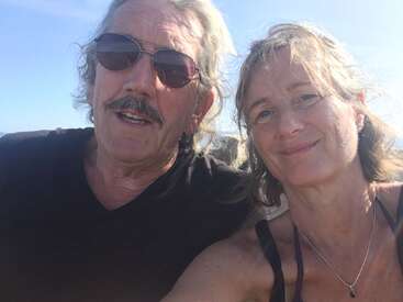 A smiling man and woman take a sunny outdoor selfie together. The man wears sunglasses and a black shirt; both look happy, relaxed, and close, enjoying the moment.