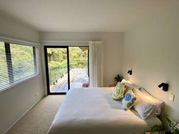 This bedroom features a large bed with colorful pillows, white walls, a sliding door to a wooden balcony, soft lighting, and large windows overlooking greenery.