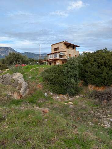 A rustic stone house sits on a grassy hill, surrounded by rocks, bushes, and mountains. The sky above is mostly cloudy, with patches of blue.
