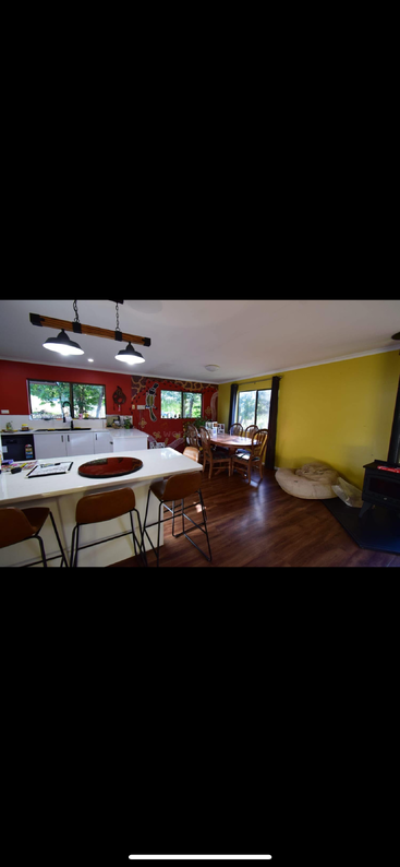 This image shows a cozy, colorful kitchen and dining area with wooden floors, yellow and red walls, modern lighting, bar stools, a dining table, and windows.
