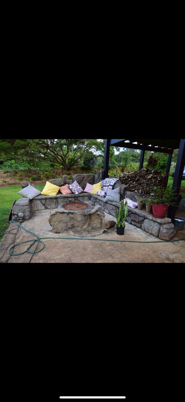 Rustic outdoor stone seating area decorated with colorful pillows, surrounded by plants, firepit in center, log pile under awning, garden hose on concrete patio, lush greenery.