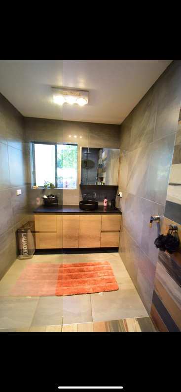 This modern bathroom features gray tiles, wooden cabinets, a black vessel sink, large mirror, laundry basket, orange rug, window, and stylish lighting, creating a cozy ambiance.