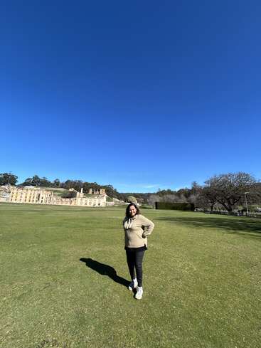 A woman stands on a large green lawn under a clear blue sky, with historic stone buildings and trees in the background, enjoying a sunny day.