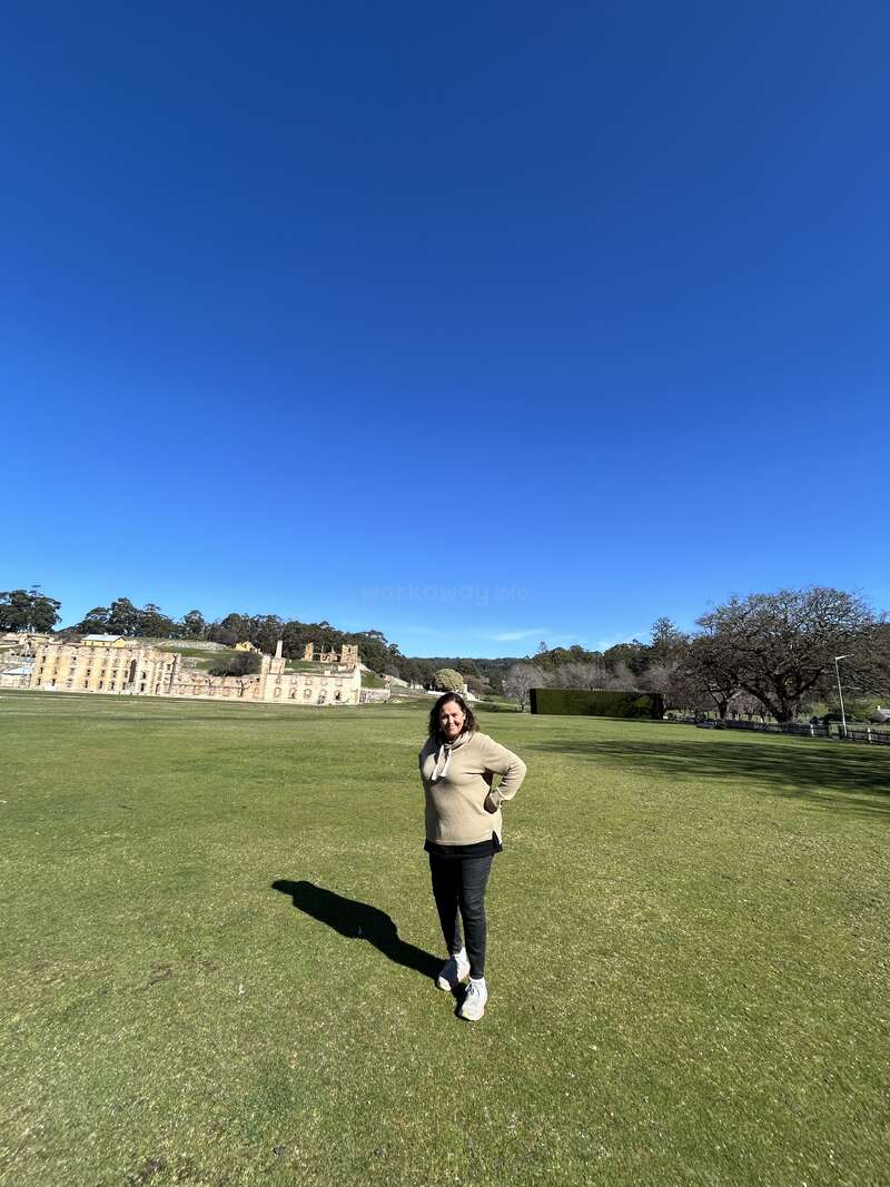 A woman stands on a large green lawn under a clear blue sky, with historic stone buildings and trees in the background, enjoying a sunny day.
