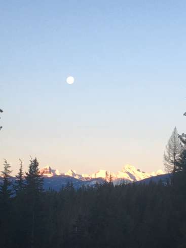 The image depicts a serene mountain range at sunset, with a full moon shining brightly in the sky above, surrounded by silhouetted trees in the foreground.