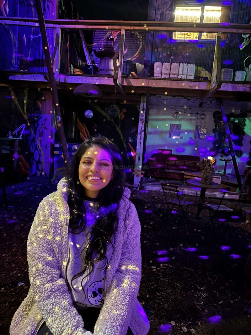 A woman with long dark hair smiles, sitting outdoors at night. Colorful lights create patterns on her fluffy coat. Background features quirky, artistic decor and seating.