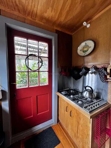 This cozy kitchen features a red door, wooden cabinets, a two-burner stove, a kettle, hanging utensils, a decorative plate, and natural light from outside.
