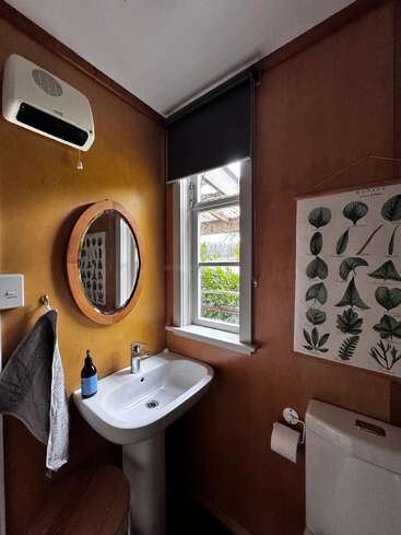 This cozy bathroom features a white sink, round mirror, towel, soap bottle, window with a view, heater, toilet, toilet paper, and botanical wall art.