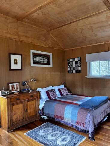 This cozy bedroom features wood-paneled walls, a double bed with colorful blankets, a vintage nightstand, eclectic wall art, a small window, and a patterned rug.