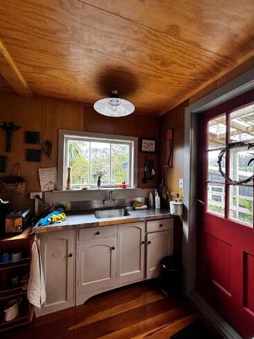 This cozy kitchen features wooden walls and ceiling, a white cabinet with a stainless steel sink, a red door, and a window overlooking a green garden.
